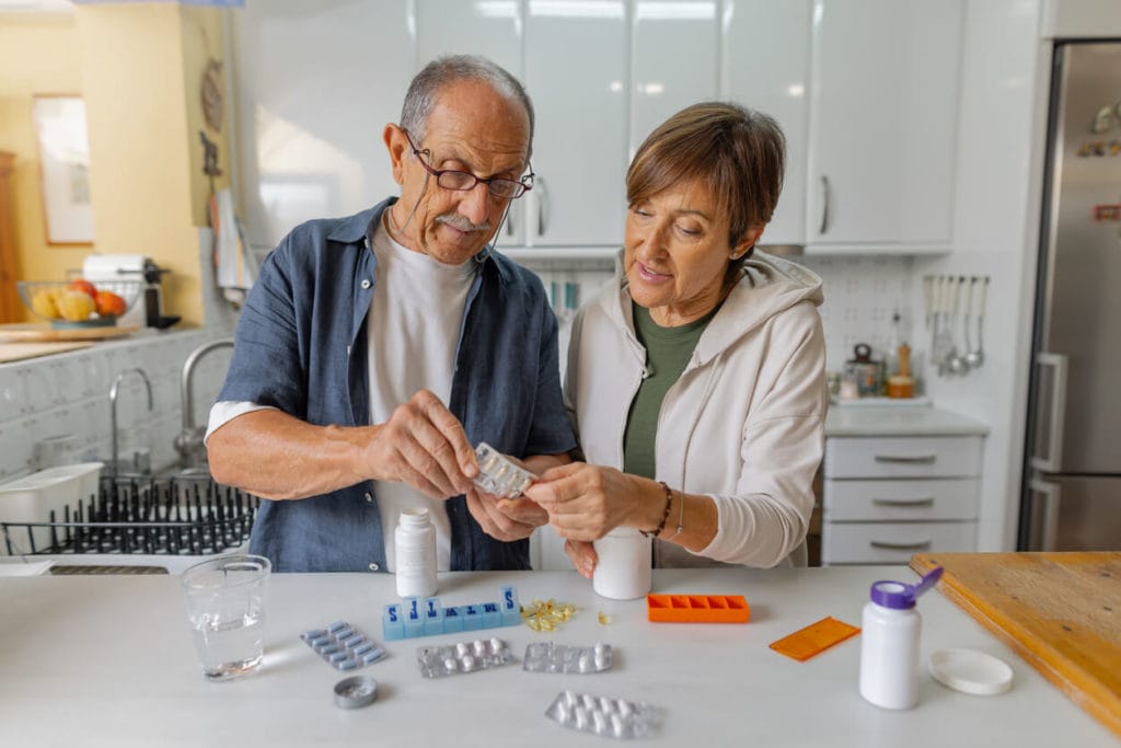 Senior medication management with a caregiver organizing a pill organizer and medication list at a kitchen table